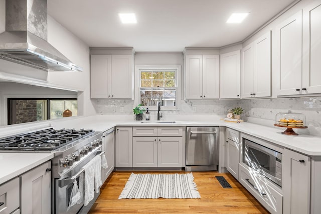 kitchen with wall chimney range hood, light hardwood / wood-style flooring, backsplash, sink, and appliances with stainless steel finishes