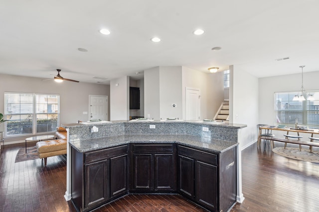 kitchen featuring ceiling fan with notable chandelier, an island with sink, dark hardwood / wood-style flooring, and pendant lighting