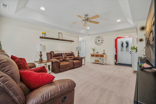 living room with a raised ceiling, ceiling fan, visible vents, arched walkways, and light carpet