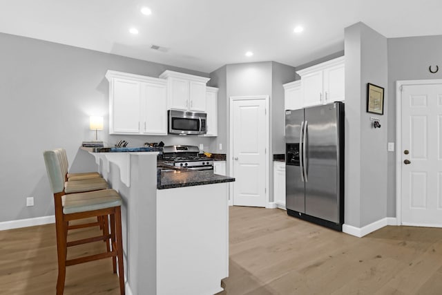 kitchen featuring light hardwood / wood-style flooring, white cabinetry, appliances with stainless steel finishes, dark stone countertops, and a kitchen bar