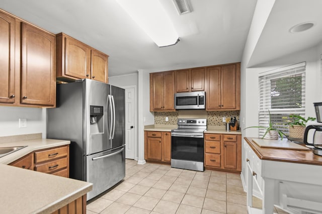 kitchen featuring stainless steel appliances, light tile patterned floors, backsplash, and brown cabinetry