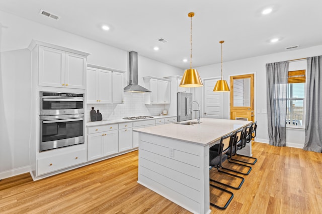 kitchen featuring hanging light fixtures, wall chimney exhaust hood, white cabinetry, stainless steel appliances, and light wood-type flooring