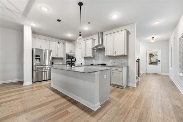 kitchen with appliances with stainless steel finishes, light wood-type flooring, wall chimney exhaust hood, a kitchen island with sink, and decorative light fixtures