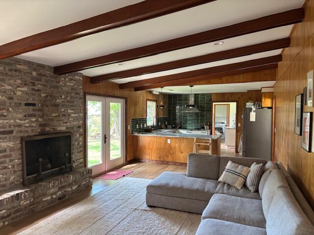 living area with french doors, a brick fireplace, beamed ceiling, light wood finished floors, and wooden walls