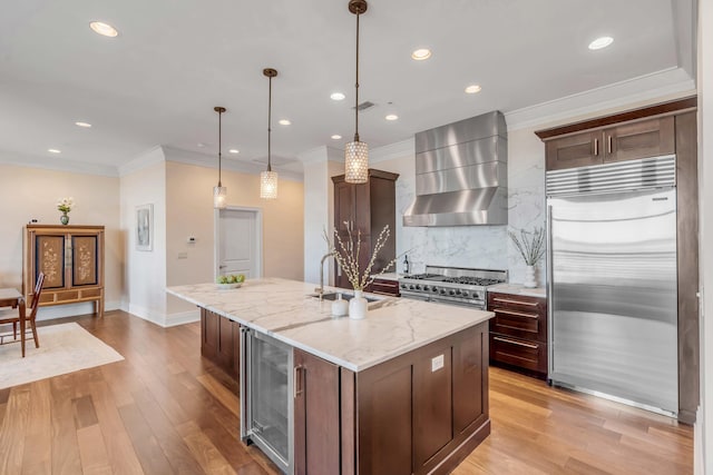 kitchen featuring high quality appliances, beverage cooler, wall chimney range hood, hanging light fixtures, and a center island with sink