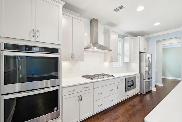 kitchen featuring wall chimney exhaust hood, dark hardwood / wood-style flooring, stainless steel appliances, ornamental molding, and white cabinetry