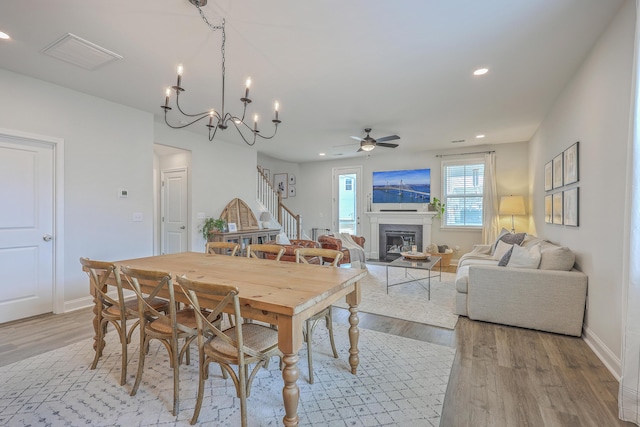 dining room featuring stairs, light wood-type flooring, recessed lighting, ceiling fan with notable chandelier, and a glass covered fireplace