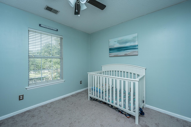 carpeted bedroom with a crib, a textured ceiling, multiple windows, and ceiling fan