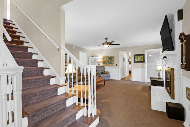 stairway featuring carpet, wainscoting, a brick fireplace, and a ceiling fan