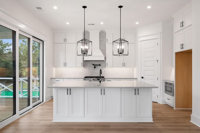 kitchen with premium range hood, an island with sink, white cabinets, decorative backsplash, and hanging light fixtures