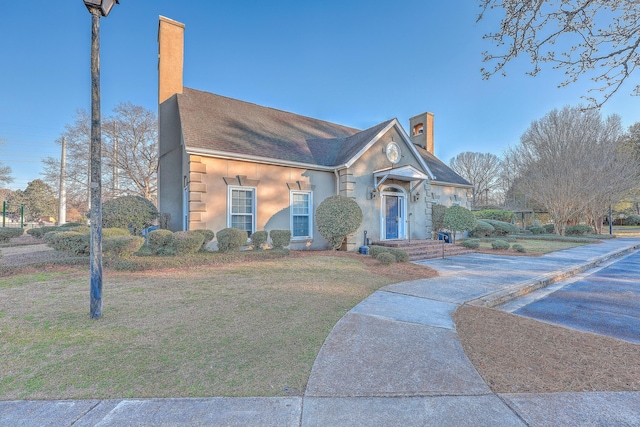 view of front facade with roof with shingles, a front lawn, a chimney, and stucco siding