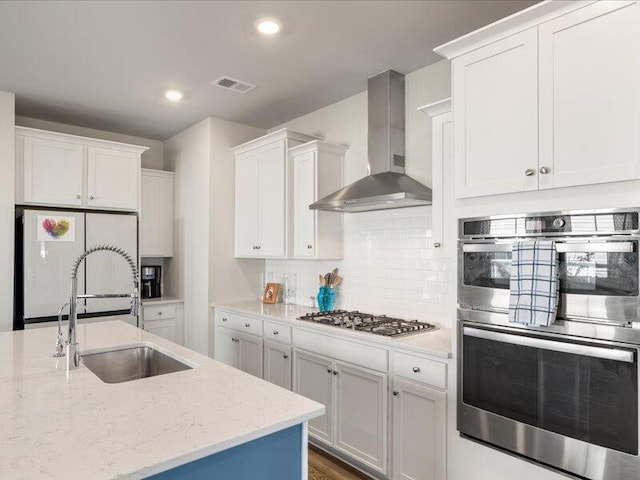 kitchen featuring white cabinetry, sink, stainless steel appliances, wall chimney range hood, and light stone counters