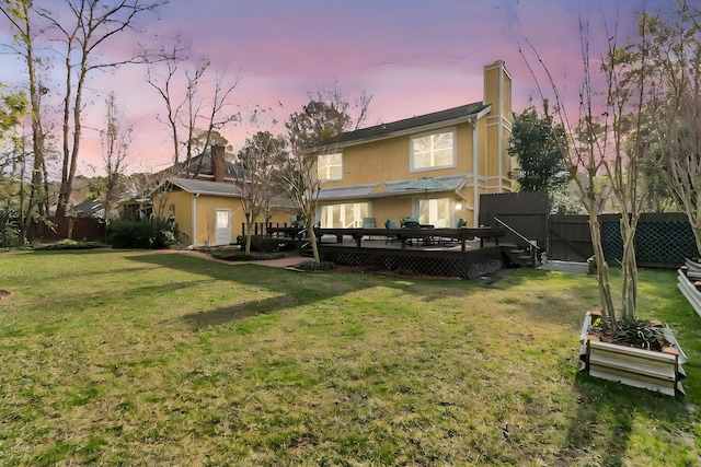 back of house at dusk featuring fence, a yard, a wooden deck, a gate, and a chimney