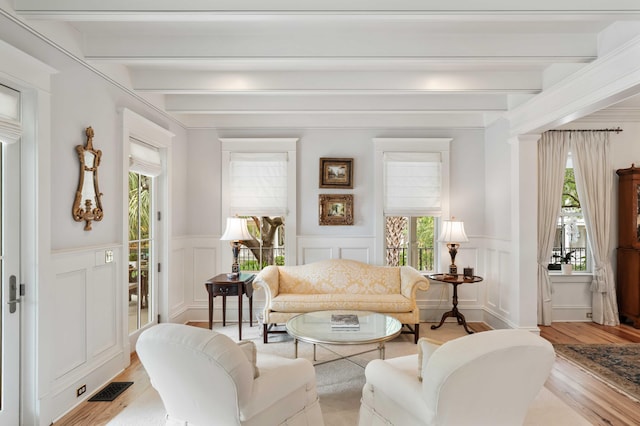 living area featuring beam ceiling, a decorative wall, light wood finished floors, and wainscoting