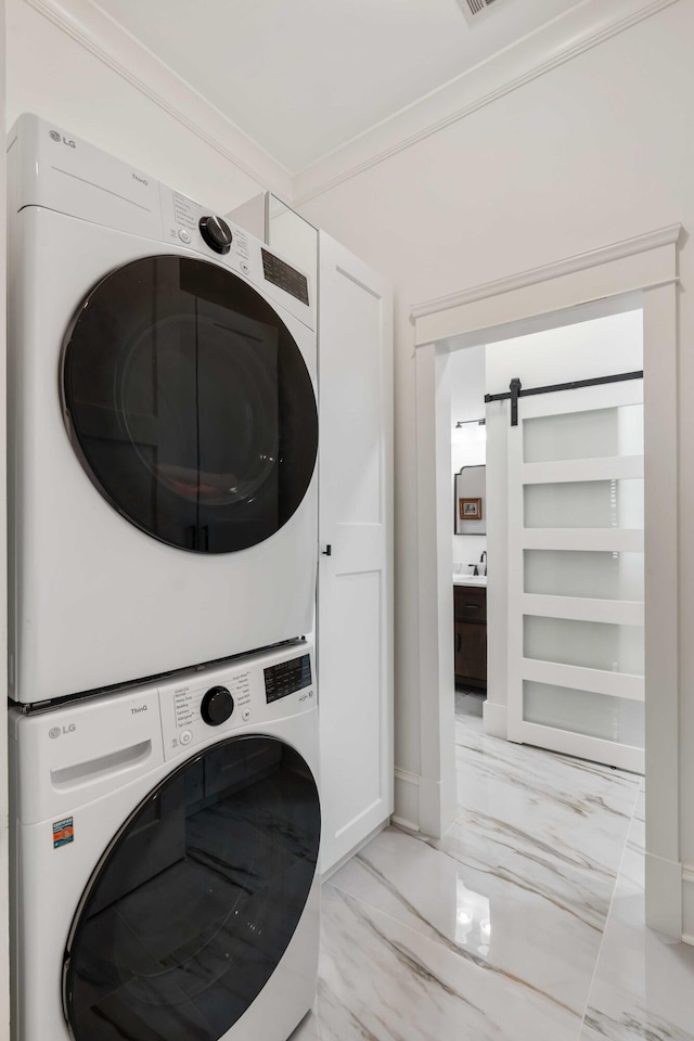 clothes washing area with stacked washing machine and dryer, light marble finish flooring, a barn door, and ornamental molding