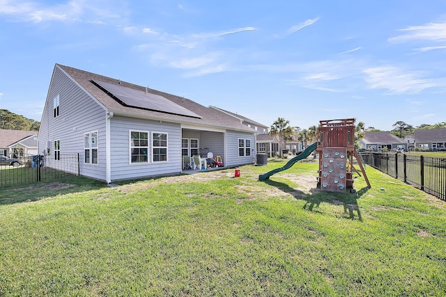 back of house featuring roof mounted solar panels, a playground, a fenced backyard, a shingled roof, and a lawn