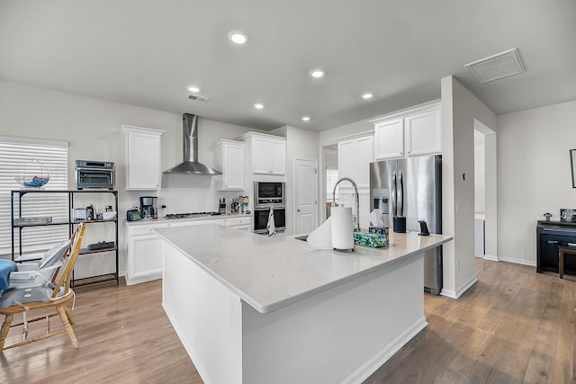 kitchen with a sink, an island with sink, light wood-type flooring, wall chimney exhaust hood, and visible vents