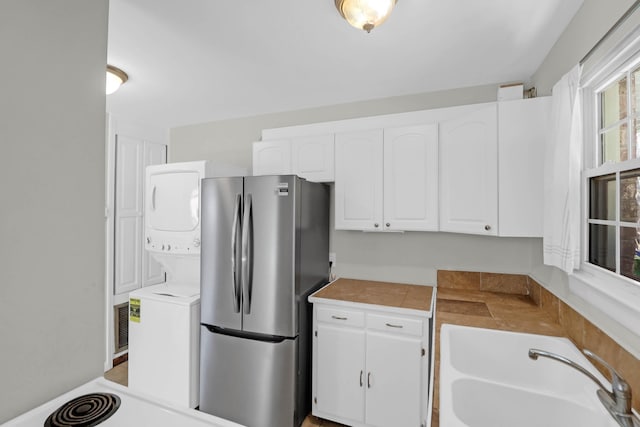 kitchen with tile countertops, freestanding refrigerator, white cabinets, a sink, and stacked washing maching and dryer