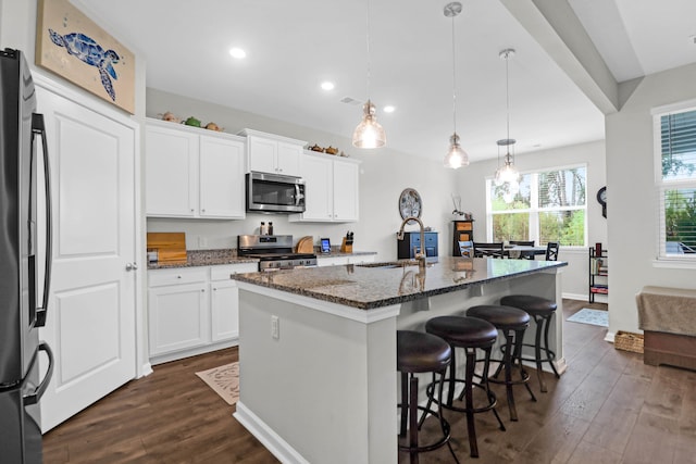 kitchen with pendant lighting, an island with sink, stainless steel appliances, dark hardwood / wood-style floors, and dark stone countertops