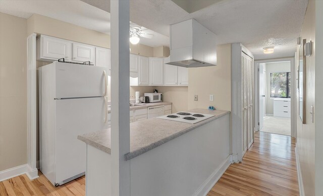 kitchen with light wood finished floors, ceiling fan, a peninsula, white appliances, and wall chimney exhaust hood