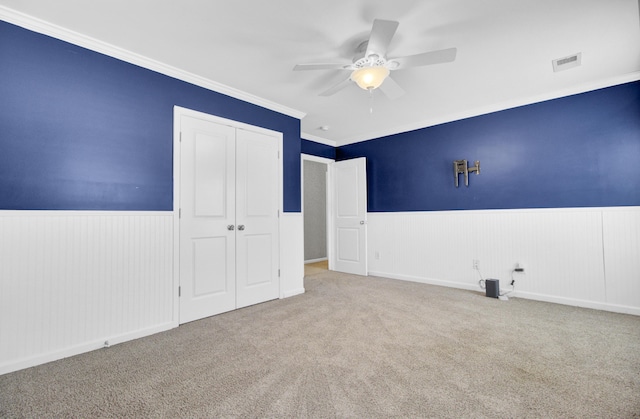 unfurnished bedroom featuring a wainscoted wall, visible vents, a closet, crown molding, and carpet flooring