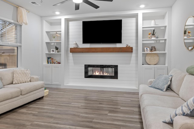 living room with a large fireplace, built in shelves, light wood-type flooring, and ceiling fan