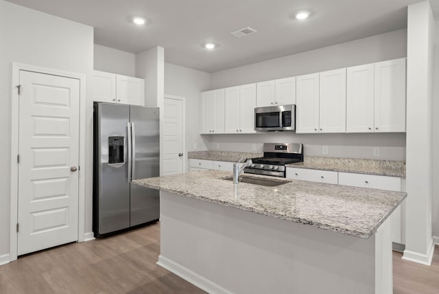 kitchen with appliances with stainless steel finishes, light stone counters, light hardwood / wood-style flooring, white cabinets, and an island with sink