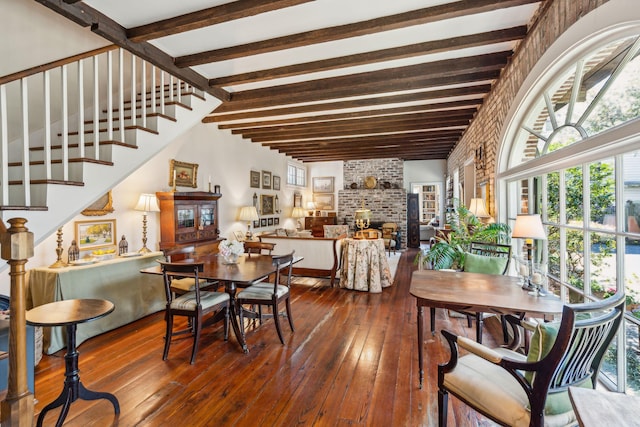 dining area featuring brick wall, a fireplace, stairs, beamed ceiling, and wood-type flooring