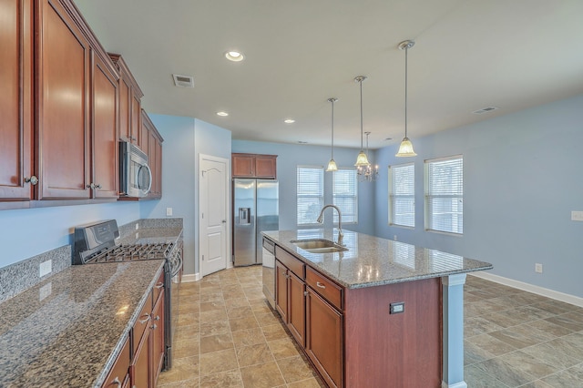 kitchen featuring a kitchen island with sink, stainless steel appliances, a sink, dark stone countertops, and decorative light fixtures