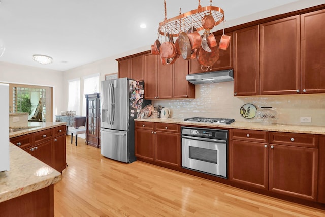 kitchen with light wood-type flooring, light stone counters, under cabinet range hood, decorative backsplash, and stainless steel appliances