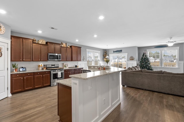 kitchen featuring ceiling fan, light stone counters, an island with sink, wood-type flooring, and appliances with stainless steel finishes