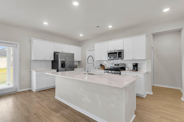 kitchen with light wood-style flooring, a kitchen island with sink, a sink, backsplash, and appliances with stainless steel finishes