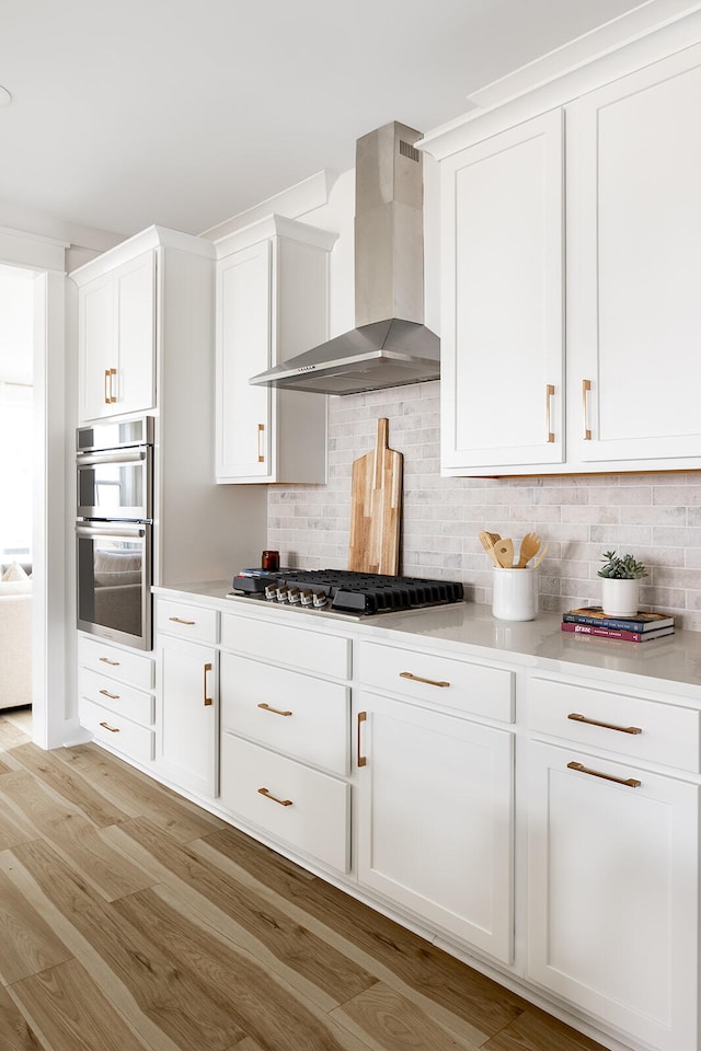 kitchen featuring white cabinets, backsplash, wall chimney range hood, appliances with stainless steel finishes, and light wood-type flooring