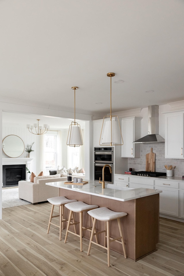 kitchen featuring decorative light fixtures, an island with sink, wall chimney exhaust hood, and light hardwood / wood-style flooring