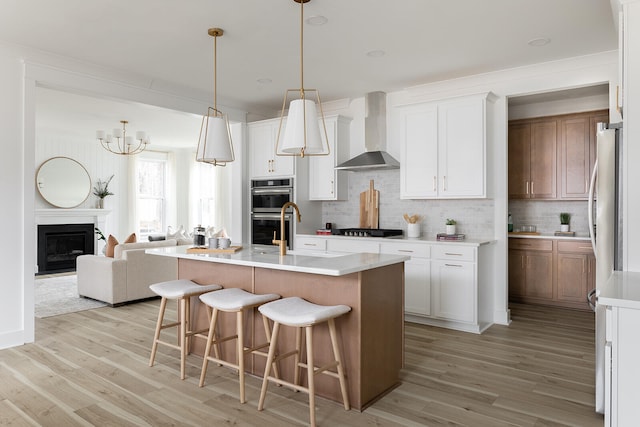 kitchen featuring white cabinets, an island with sink, wall chimney exhaust hood, light hardwood / wood-style flooring, and stainless steel appliances