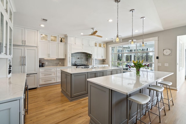 kitchen with light hardwood / wood-style flooring, a kitchen island with sink, hanging light fixtures, gray cabinetry, and tasteful backsplash