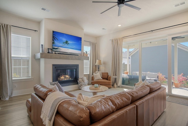 living area with light wood-type flooring, visible vents, a glass covered fireplace, and a ceiling fan