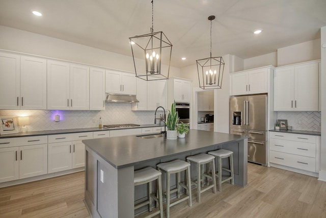 kitchen with a center island with sink, under cabinet range hood, a sink, dark countertops, and stainless steel appliances