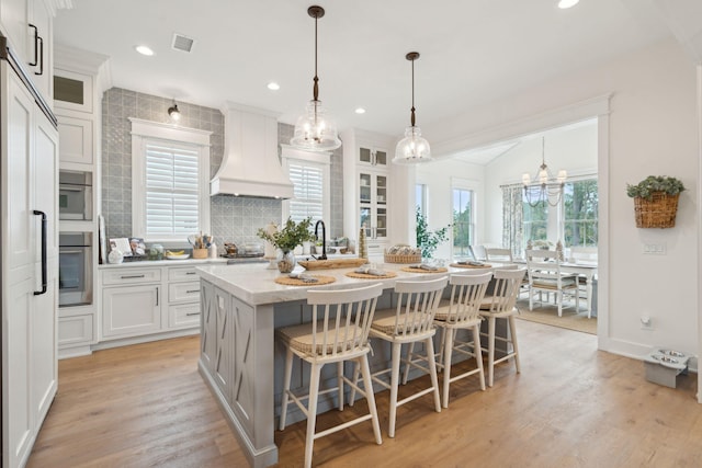 kitchen with custom exhaust hood, white cabinetry, hanging light fixtures, and a center island with sink