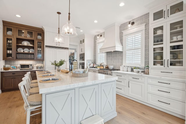 kitchen featuring a kitchen bar, custom exhaust hood, decorative light fixtures, an island with sink, and white cabinets