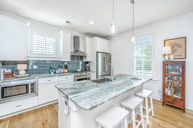 kitchen with wall chimney exhaust hood, an island with sink, light wood-type flooring, and premium appliances