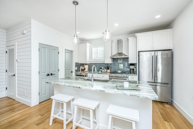 kitchen featuring stainless steel appliances, a kitchen island with sink, sink, wall chimney range hood, and white cabinetry