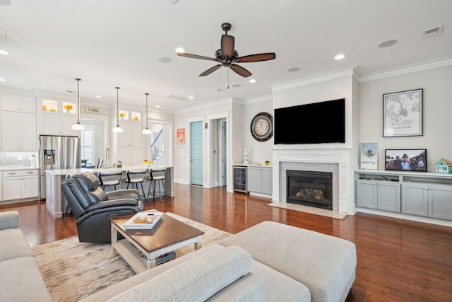 living room featuring beverage cooler, a fireplace with flush hearth, ornamental molding, dark wood-style flooring, and recessed lighting