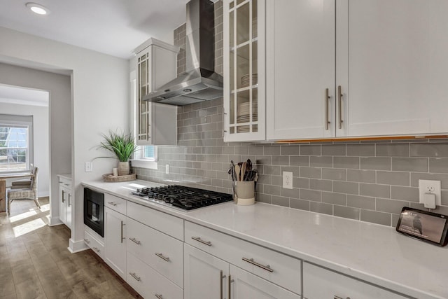 kitchen featuring backsplash, glass insert cabinets, dark wood-type flooring, wall chimney range hood, and stainless steel gas stovetop