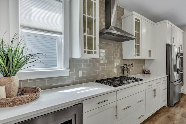 kitchen featuring stainless steel appliances, white cabinets, glass insert cabinets, wall chimney range hood, and tasteful backsplash