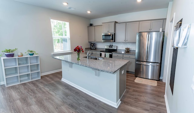 kitchen with light stone countertops, stainless steel appliances, sink, gray cabinets, and an island with sink