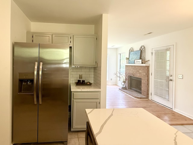 kitchen with stainless steel fridge with ice dispenser, a brick fireplace, light wood-style floors, baseboards, and tasteful backsplash