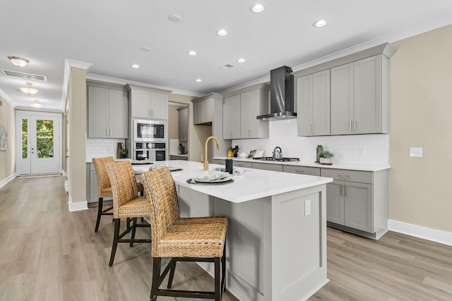 kitchen with gray cabinets, a kitchen island with sink, a breakfast bar area, wall chimney exhaust hood, and appliances with stainless steel finishes