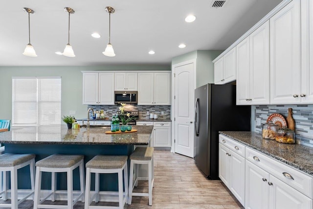 kitchen with a kitchen island with sink, backsplash, white cabinetry, dark stone counters, and appliances with stainless steel finishes