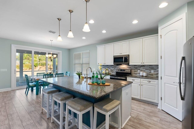 kitchen featuring decorative light fixtures, white cabinets, dark stone countertops, a center island with sink, and appliances with stainless steel finishes